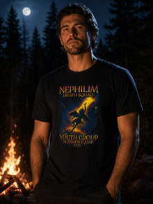 Man wearing the black Nephilim Death Squad Youth Group Summer Camp 2025 T-shirt beside a campfire in a pine forest at night