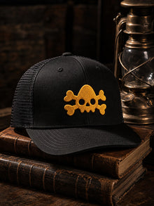 Black Nephilim Death Squad logo trucker hat with the gold NDS skull and crossbones, styled on antique books beside a brass lantern