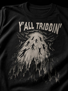 Close-up of the cream Y'all Tribbin' rapture print on a black T-shirt: beam of light lifting people above the city